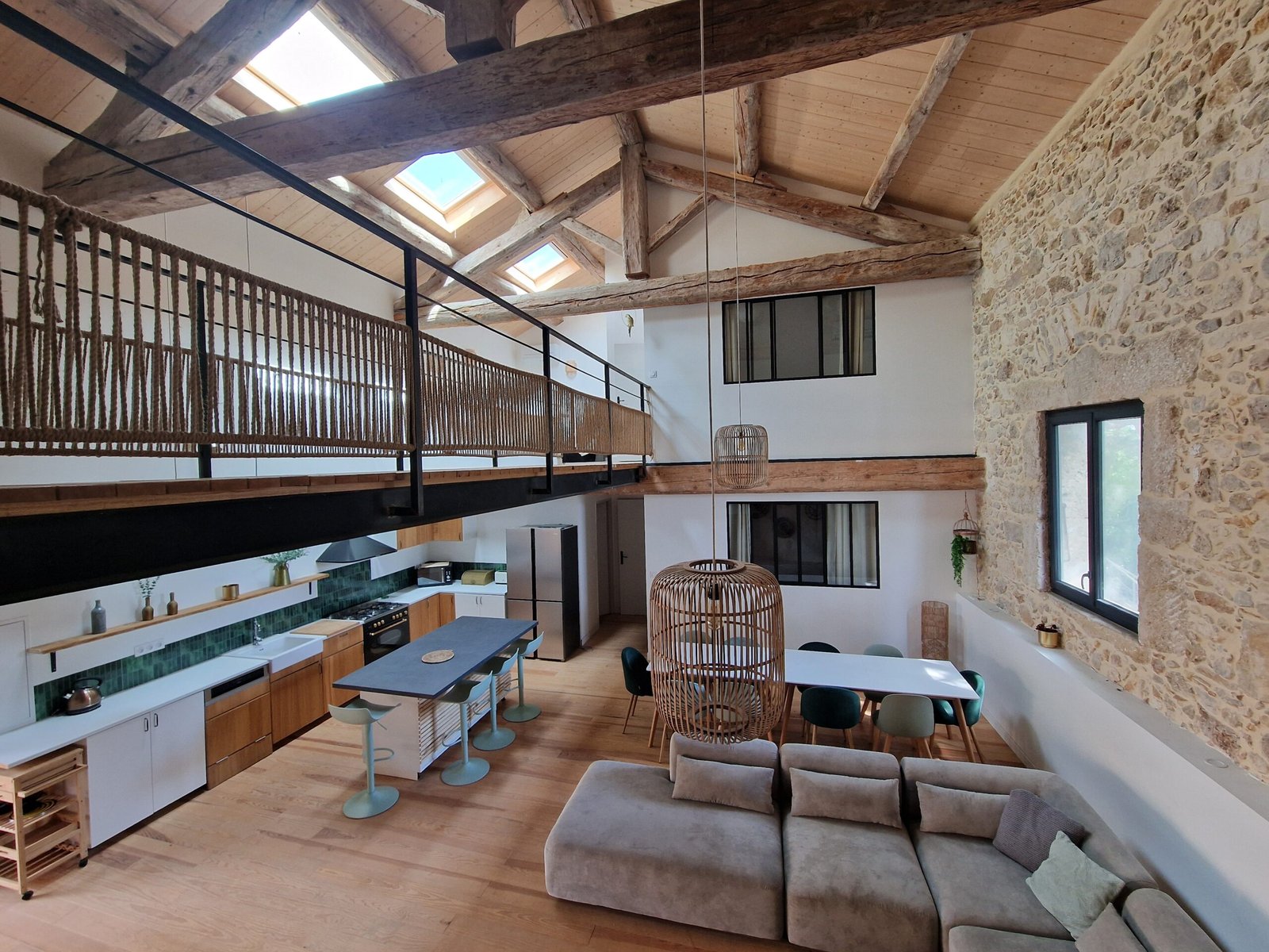 Grand séjour du gîte à Narbonne avec cuisine ouverte, grande table, salon, mur en pierre, charpente apparente et passerelle en bois.