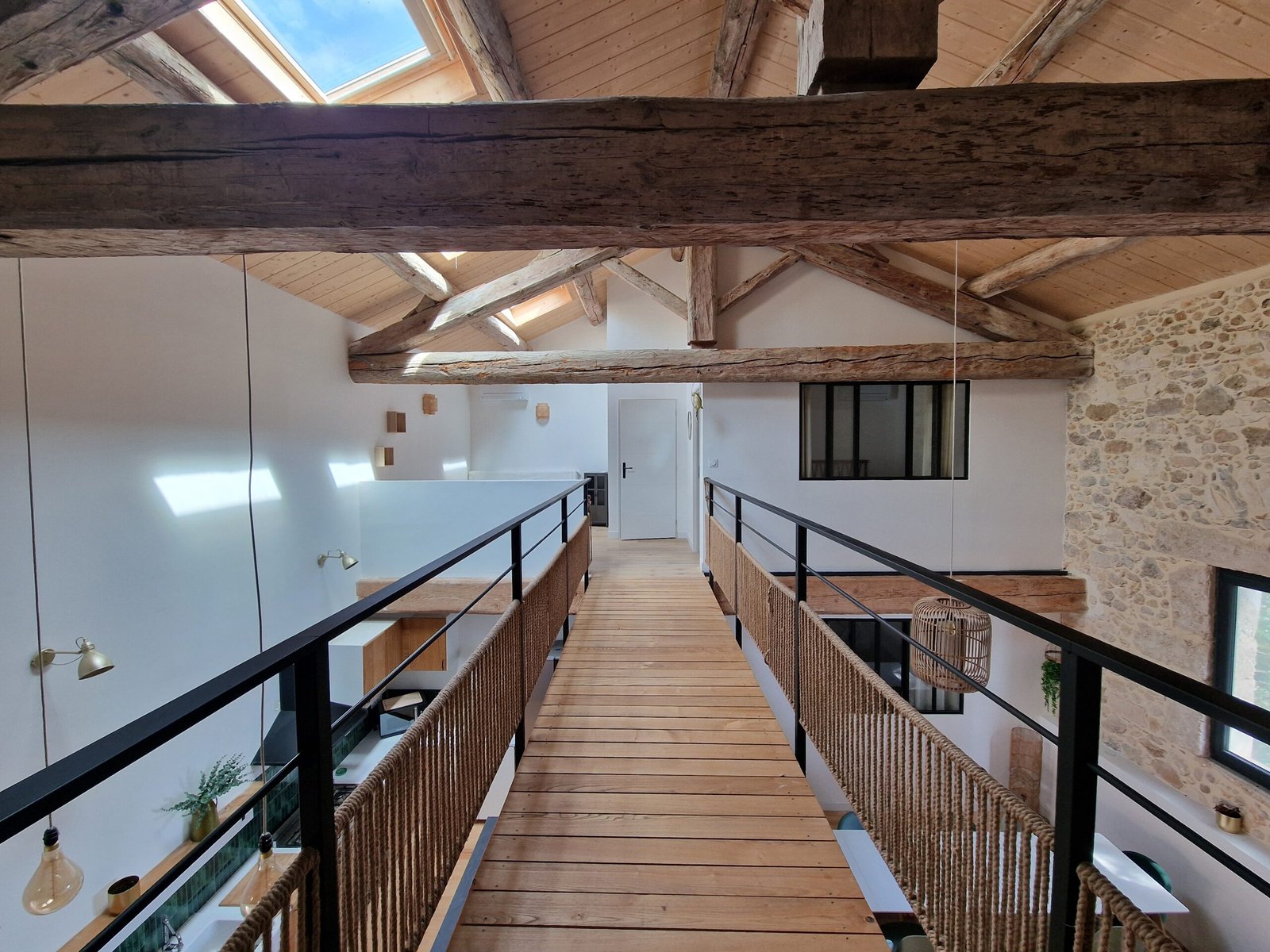 Passerelle intérieure en bois du gîte à Narbonne, entourée d'une charpente apparente et d'un grand mur en pierre.